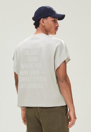 Muž v svetlobéžovom oversized tričku s vybledlým nápisom na chrbte, olivovo zelených nohaviciach a námorníckom modrom šiltovke, otočený do strán.