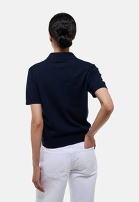 Polo-shirt marine à manches courtes, col et ourlet côtelés, présenté de dos, associé à un pantalon blanc. Texture lisse sans motifs.