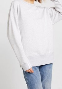 Šedá bavlněná mikina s dlouhými rukávy, žebrovanými manžetami a páskem na spodním lemu. Na hrudi je vyšívané logo a kontrastní bílé pruhy.