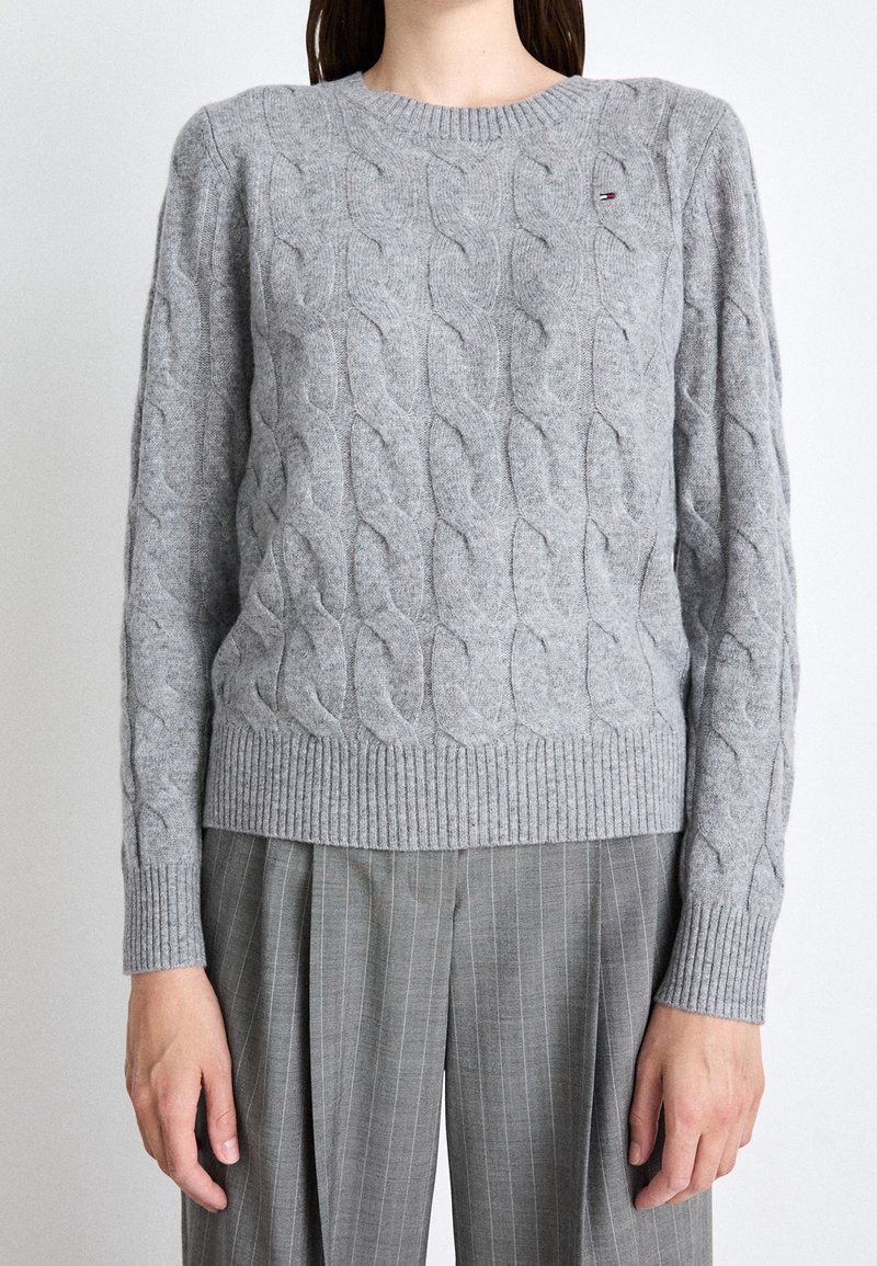 Femme portant un pull en maille torsadée gris clair avec des poignets et un ourlet côtelés, assorti à un pantalon plissé gris à fines rayures.