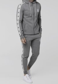 Szary komplet bluzy i spodni z miękkiego materiału, z białym logo i paskami na rękawach oraz nogawkach. Do tego białe trampki.