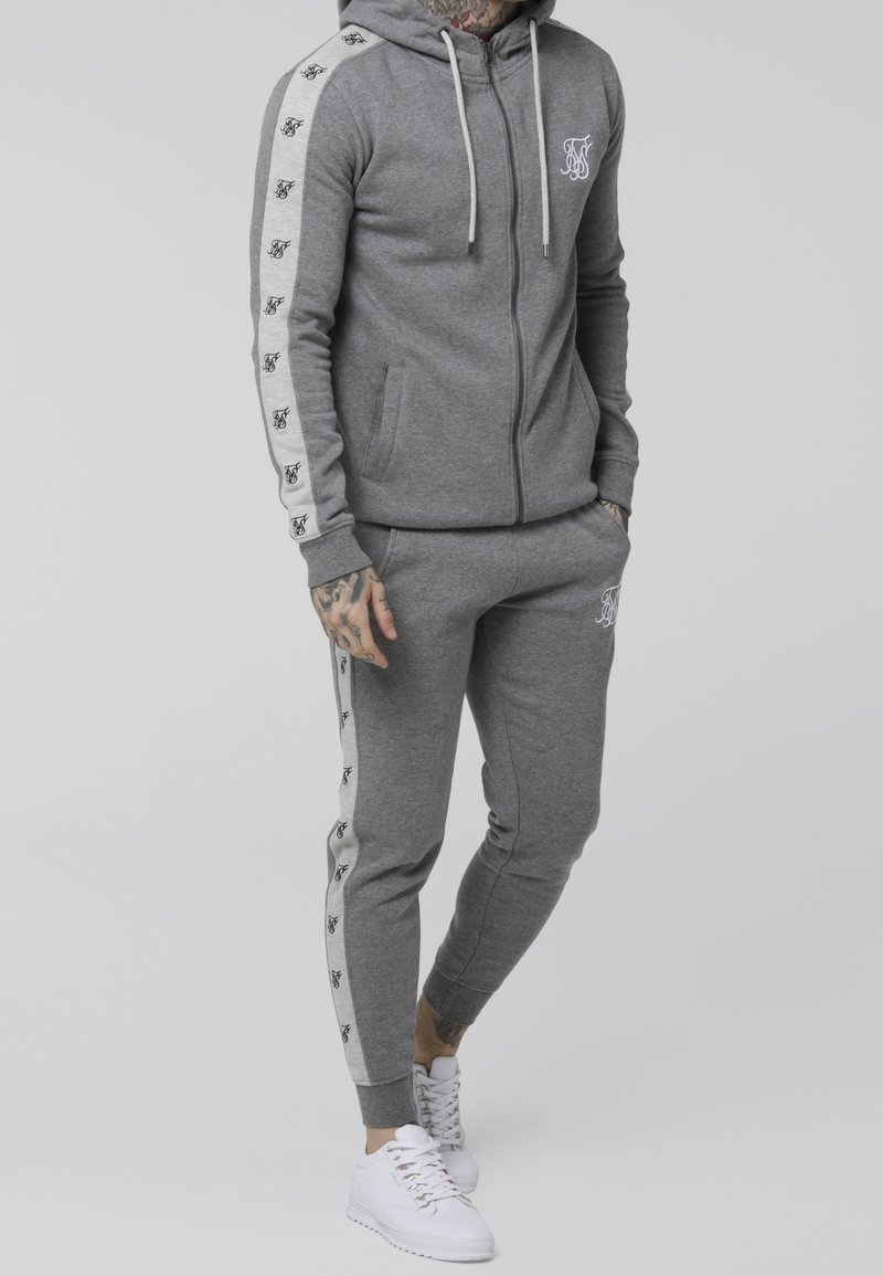 Szary komplet bluzy i spodni z miękkiego materiału, z białym logo i paskami na rękawach oraz nogawkach. Do tego białe trampki.