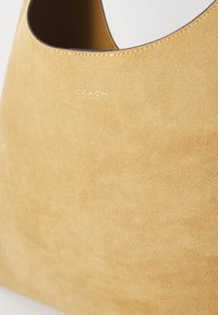 Hobo bag in beige suede with smooth texture, featuring a round opening and subtle gold "COACH" logo. Simple design without embellishments.