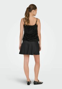 Haut noir sans manches avec franges texturées, associé à une jupe noire ajustée. Le mannequin porte des chaussures noires cloutées.