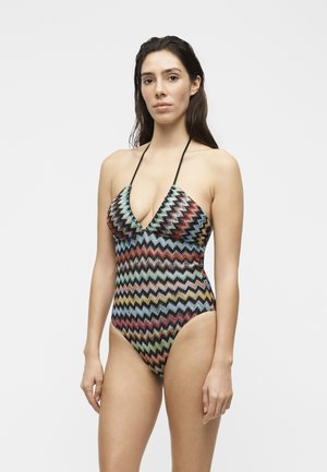Femme aux longs cheveux foncés portant un maillot de bain une-pièce à col halter avec un motif zigzag multicolore, sur un fond clair uni.