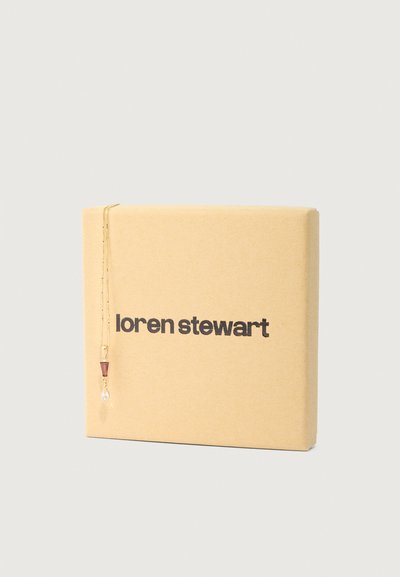 Zlatna ogrlica s crvenim trokutaškim privjeskom i prozirnom kapljicom, obješena na smeđoj kraft kutiji s natpisom "loren stewart" u podebljanom crnom tekstu.