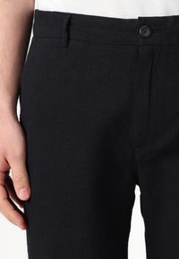 Black linen trousers with a textured finish, featuring a button closure and a slim fit. Less than full length, with side pockets visible.