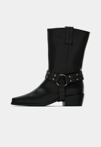 Μαύρες δερμάτινες μπότες μέχρι το γόνατο με μυτερή μύτη, διακοσμημένες με ζώνη με καρφιά και με chunky τακούνι για επιπλέον σταθερότητα.