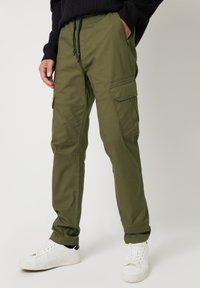 Groene cargobroek van stevige stof, met meerdere zakken, een trekkoord in de taille en taps toelopende benen. Gecombineerd met witte sneakers.