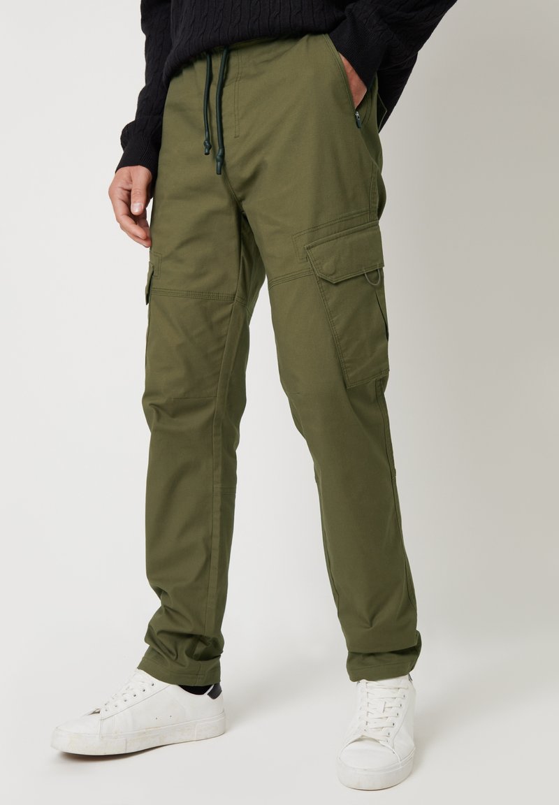 Groene cargobroek van stevige stof, met meerdere zakken, een trekkoord in de taille en taps toelopende benen. Gecombineerd met witte sneakers.