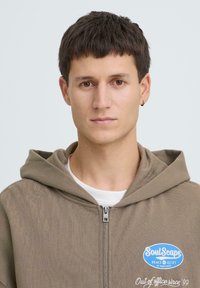 Sweat à capuche marron avec fermeture éclair, en tissu texturé, arborant un patch logo bleu et blanc. Comprend une capuche et une fermeture éclair apparente.