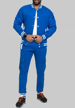 Survêtement de sport bleu avec des accents rayés blancs, comprenant une veste bomber zippée et un pantalon cargo avec des poches latérales.