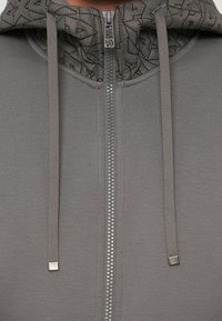 Felpa grigia con zip e cappuccio trapuntato. Presenta cordini e una cerniera argentata. Tessuto liscio con un sottile motivo geometrico.