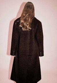 Ruda vilnos striukė su ilga, tiesia forma. Savybėmis yra mygtukų apykaklės ir lygi tekstūra, matoma iš nugaros prieš rožinę fono.