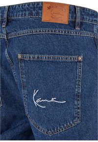 Mørkeblå denimjeans med lærpatch som har "Karl Kani"-logo, hvit brodert signatur på baklommen, synlige sømdetaljer.
