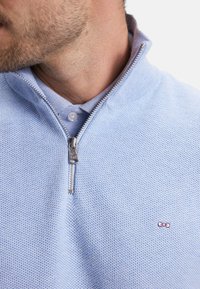 Pull-over à col zippé bleu clair en tissu texturé, avec une fermeture éclair argentée et un petit logo brodé violet sur la poitrine gauche.