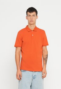 Oranssi polo-paita, joka on valmistettu puuvillasta, klassisella kauluksella, lyhyillä hihoilla ja kolmella napilla kaula-aukolla.