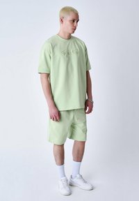 T-shirt en coton vert clair à manches courtes avec logo brodé, assorti à un short. Porté avec des baskets blanches et des chaussettes jusqu'à la cheville.