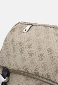 Sac à dos en tissu beige avec un motif de monogramme répété, tirette en métal argenté, et une sangle noire avec un détail de boucle argentée.