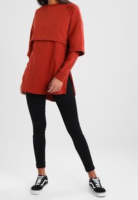 Sudadera roja de gran tamaño con un superpuesto recortado, aberturas laterales y detalle de texto en relieve; combinada con jeans ajustados negros y zapatillas negras.