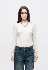 Top de manga comprida, bege claro com um padrão subtil, combinado com jeans azuis escuros de corte largo e um colar de corrente prateada.