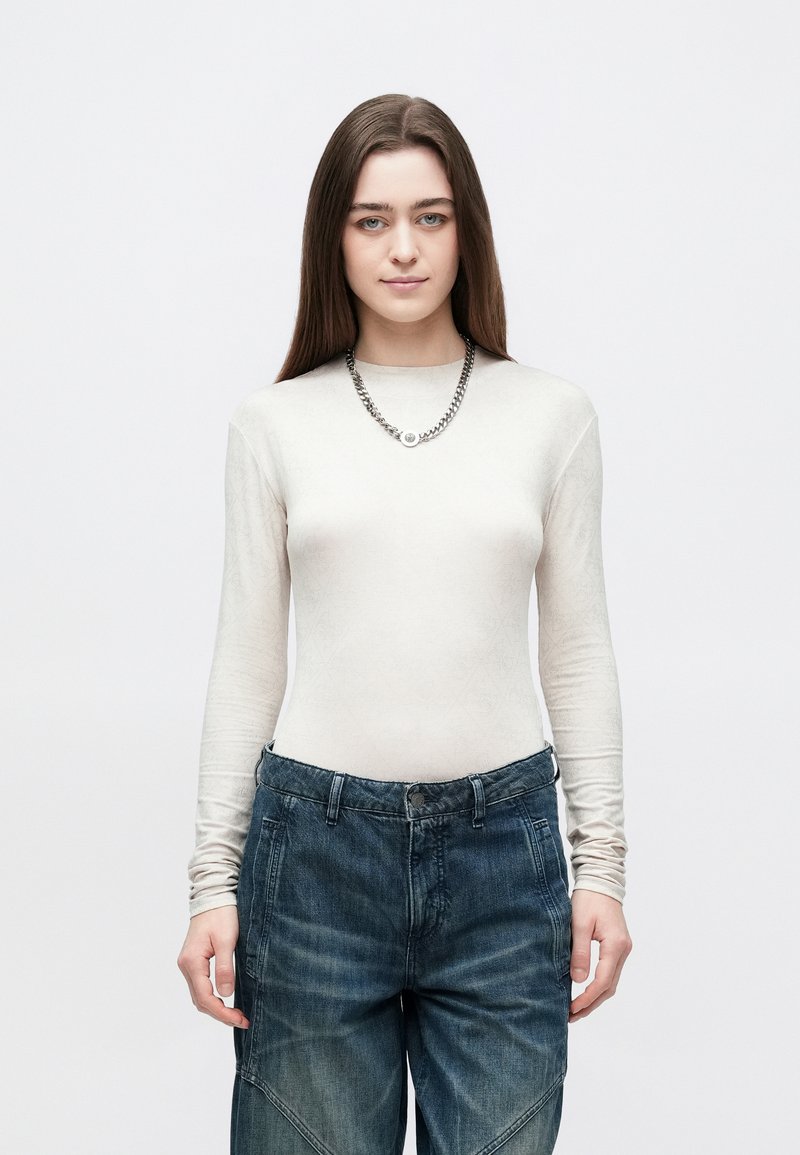 Top de manga comprida, bege claro com um padrão subtil, combinado com jeans azuis escuros de corte largo e um colar de corrente prateada.