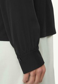 Schwarze Bluse mit langen Ärmeln und gerafften Ärmeln, ausgestattet mit Knopfmanschetten und einer glatten Textur. Taillierter, lockerer Schnitt mit filigranen Details.