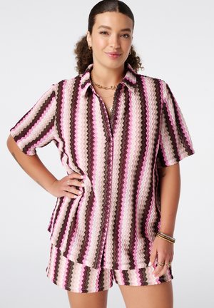 Femme aux cheveux bouclés portant un t-shirt à manches courtes à motif zigzag rose, marron et blanc, et un short assorti, accessoirisés avec des bijoux en or.