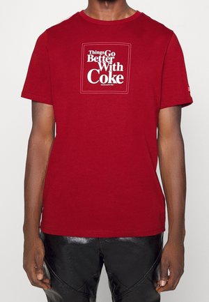 Osoba ubrana w czerwoną koszulkę z białym napisem "Things Go Better With Coke" i czarne błyszczące spodnie na jednolitym tle.