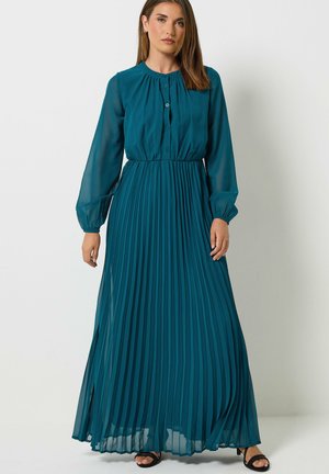 Femme portant une robe longue plissée à manches longues couleur sarcelle, debout devant un fond blanc uni, chaussée de sandales noires à bouts ouverts.