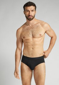 Zwarte herenonderbroeken van gladde stof, met een lage taille en een elastische tailleband. Het model heeft kort, donker haar en een baard.