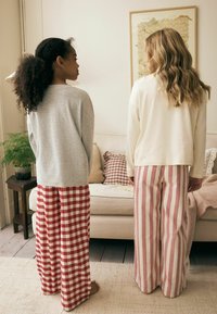 Grauer strukturierter Pullover, rot karierte weit geschnittene Hose. Weißes geripptes Oberteil, rosa gestreifte weit geschnittene Hose. Schlichtes Wohnzimmer mit Kissen.