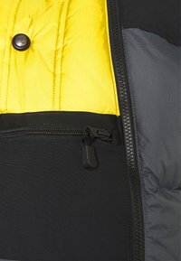 Puchowa kurtka z żółtym wnętrzem, czarną zewnętrzną stroną, kieszenią na zamek i trwałymi materiałami o teksturowanym wykończeniu.