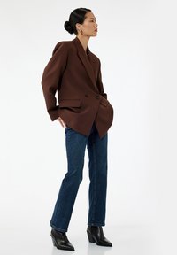 Blazer marrone doppiopetto con larghe rever, abbinato a jeans aderenti blu scuro e stivaletti neri con tacco. Tessuto liscio e strutturato.