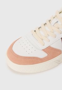 Fehér sneaker rózsaszín velúr részletekkel, kerek orral, perforált díszítéssel, krémszínű fűzőkkel és texturált gumitalppal, amely geometrikus mintázattal rendelkezik.