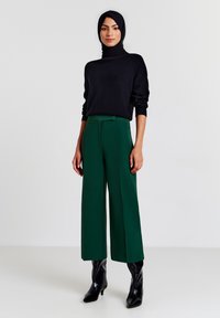 Maglione nero a collo alto, pantaloni a gamba larga verdi e stivali neri fino al ginocchio. Tessuto a maglia spessa, design strutturato e texture liscia.