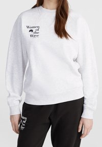 Mujer vistiendo un sudadera gris claro y pantalones negros, ambos con el texto "Women of the Wave" impreso en la parte superior izquierda.