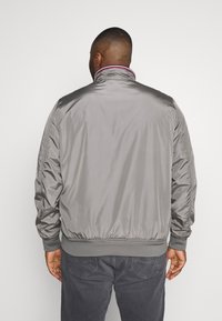 Man wearing a light gray jacket with ribbed cuffs and collar, standing facing away against a plain white background.