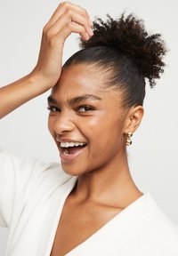 Persona sonriente con cabello rizado recogido en un moño, usando pendientes de aro dorados. Tono de piel claro, vistiendo una prenda blanca texturizada. Fondo neutro.