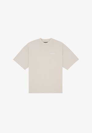 Kobieta o ciemnych kręconych włosach, ubrana w oversize beżowy T-shirt oraz pastelowo różowe i niebieskie szorty w paski, na jednolitym tle.