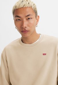 Beige sweatshirt met een ronde hals, zachte textuur en een rode Levi's logo patch op de linkerkant van de borst. Heeft een relaxte pasvorm en ribgebreide boorden.