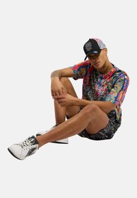Fargerik grafisk t-skjorte med abstrakte mønstre, svart caps med broderte detaljer, mønstrede shorts og hvite joggesko med svarte detaljer.