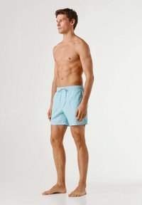 Jeune homme debout, pieds nus, portant un short de bain bleu clair à motifs, regardant sur le côté sur un fond blanc uni.