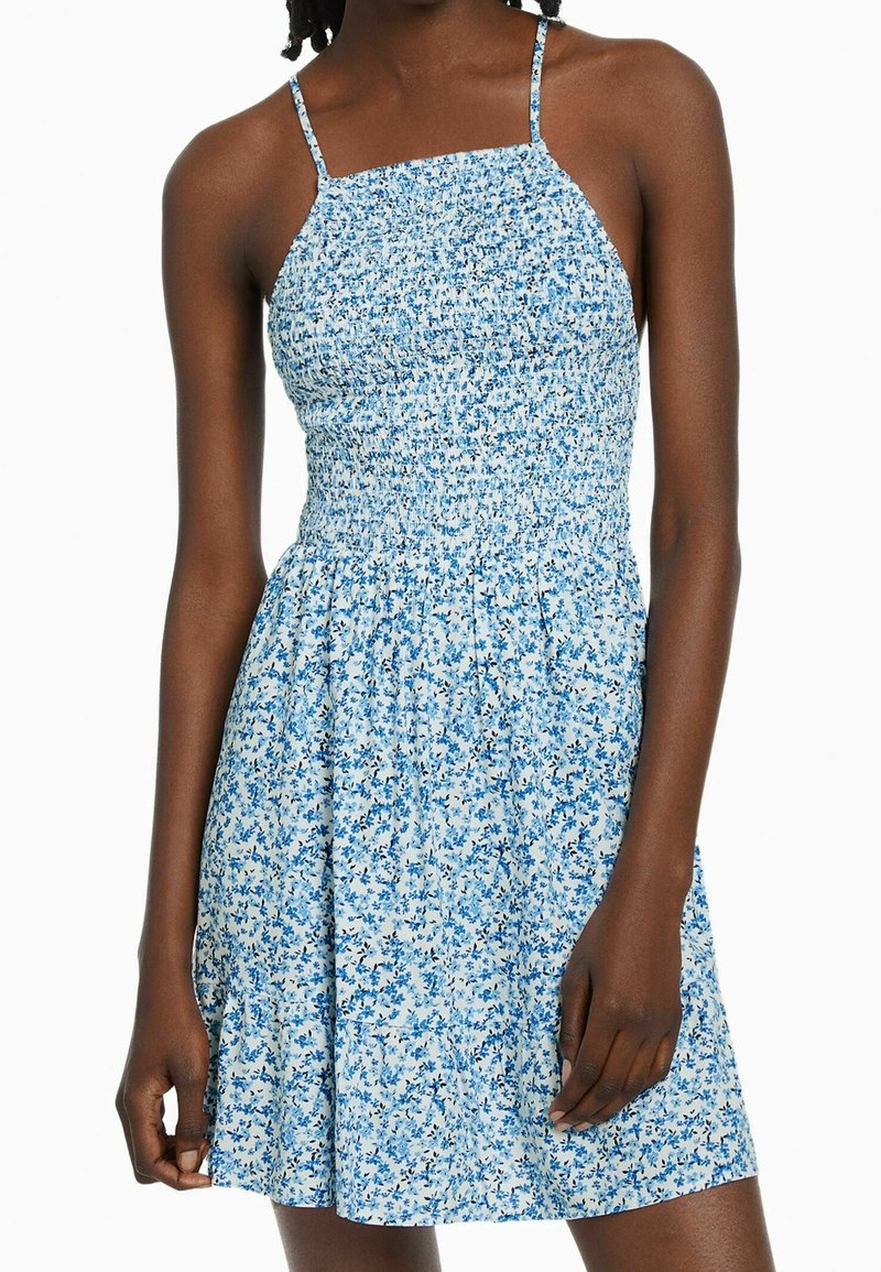 Femme portant une robe d'été sans manches, bleue et blanche à motifs floraux, avec de fines bretelles et un corsage smocké ajusté.