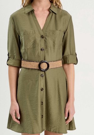 Femme portant une robe boutonnée vert olive avec manches retroussées et une large ceinture tissée avec une grande boucle ronde noire.
