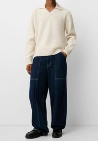Personne portant un pull crème à col en V, un pantalon en denim bleu foncé à jambes larges avec coutures visibles, et des mocassins noirs, debout avec une main dans la poche.