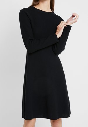 Vrouw draagt een zwarte, geribbelde, gebreide jurk met lange mouwen en een ronde hals en past de mouw aan tegen een effen lichtgrijze achtergrond.