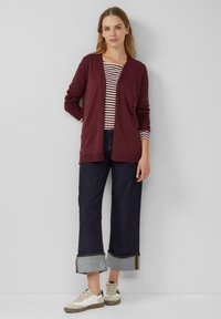 Burgunderfarbener Cardigan mit langen Ärmeln und offenem Stil über einem gestreiften Oberteil. Dunkelblaue Jeans im weitem Bein mit kontrastierenden Säumen und hellen Sneakers.