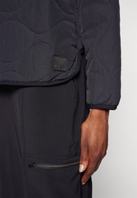 Zwarte gewatteerde jas met een textuurafwerking, voorzien van een logopatch en een elastische manchette. Gecombineerd met zwarte, aansluitende broek met een ritszak.