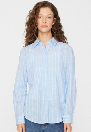 Frau mit langen lockigen Haaren, die ein hellblau-weiß vertikal gestreiftes Hemd und blaue Jeans trägt, steht vor einem einfarbigen Hintergrund.